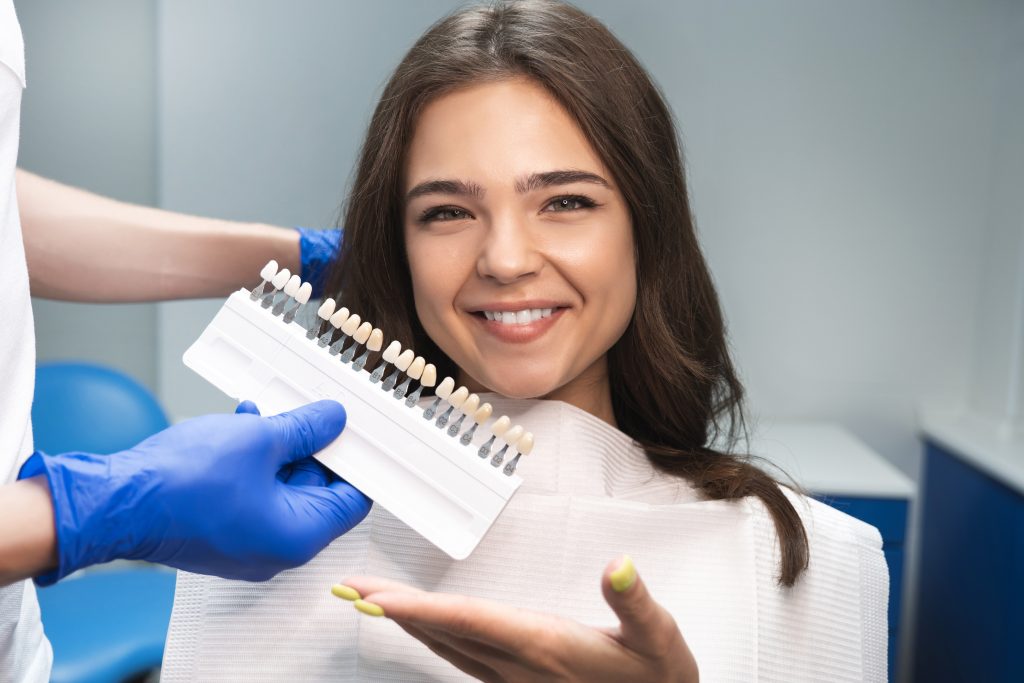smiling brunette woman patient having appiontment in dental clinic picking up shade using tooth enamel scale held by dentist in blue gloves.